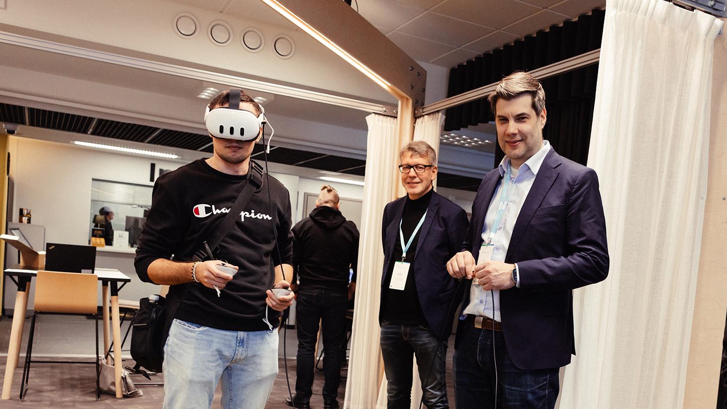 Image for Joint Education Lab Projects Call where one person is using VR-glasses and two persons guiding him.